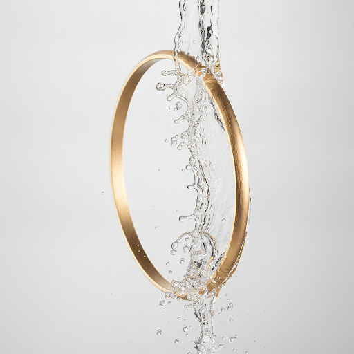 Close-up product photography of a sleek, minimalist gold bangle with a brushed finish, submerged in clean, splashing water to demonstrate water resistance. High-speed photography, crisp droplets, elegant lighting. Soft off-white background. Editorial luxury jewelry style.